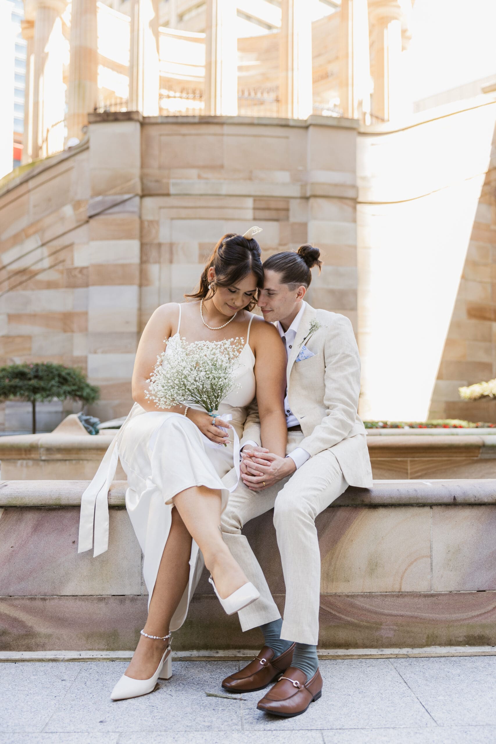 Bride and groom sitting closely together during a Brisbane city elopement, holding hands in soft afternoon light with the bride holding a bouquet of baby’s breath.