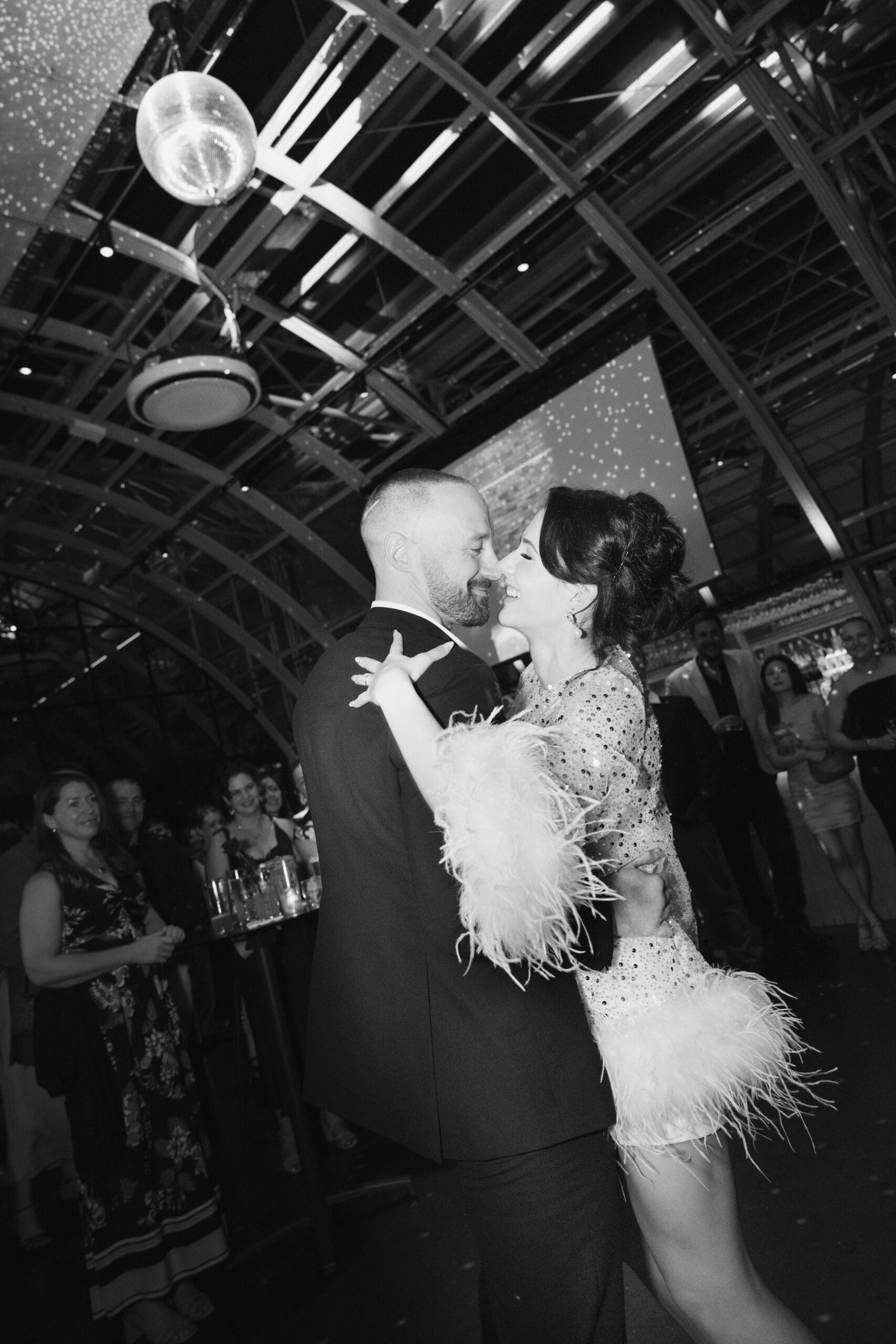 Bride and groom sharing their first dance at their wedding reception, surrounded by guests in a dimly lit venue, captured in a candid black and white moment.