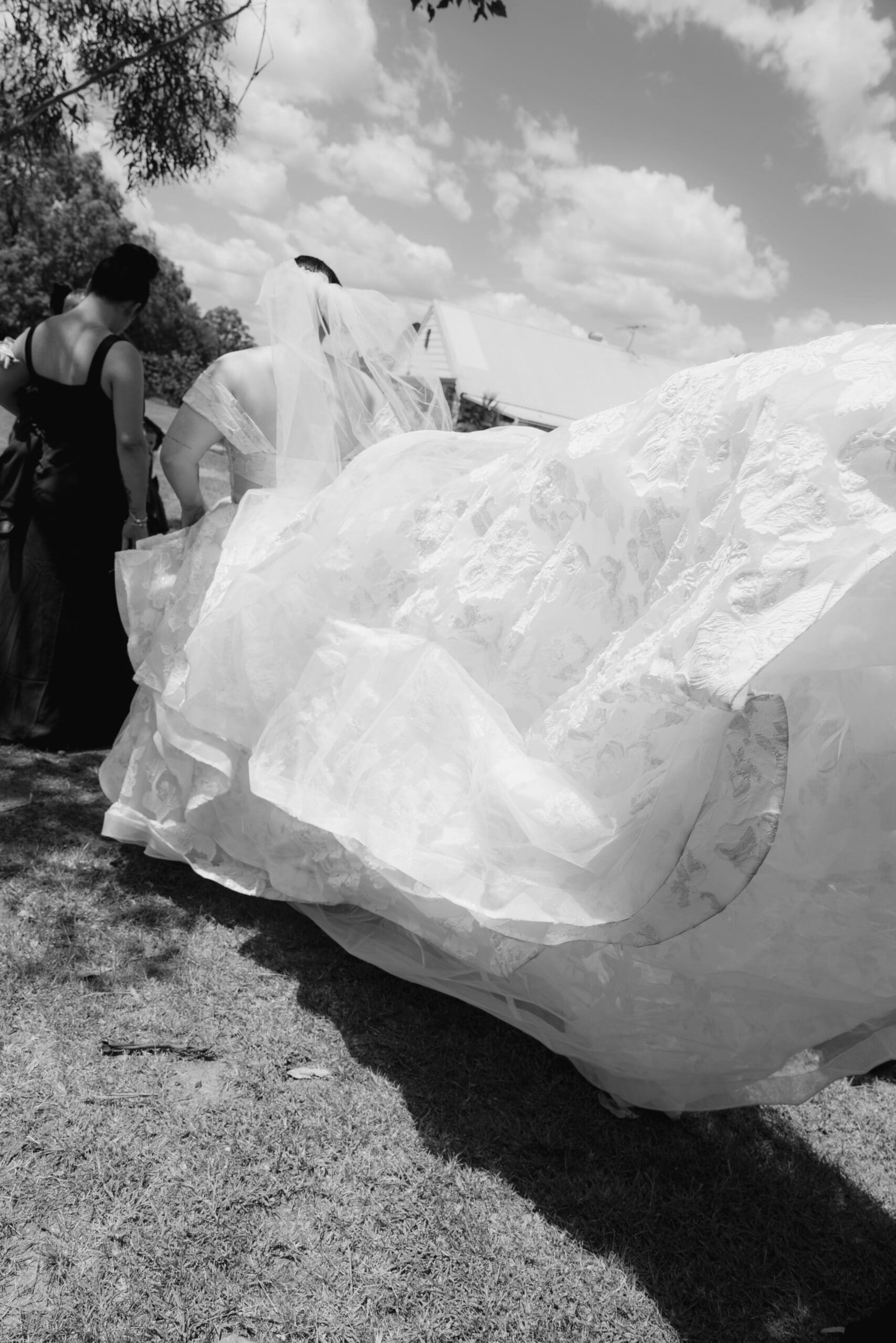 Bride walking outdoors on her wedding day while bridesmaids help carry the flowing train of her gown, captured in a candid black and white moment.