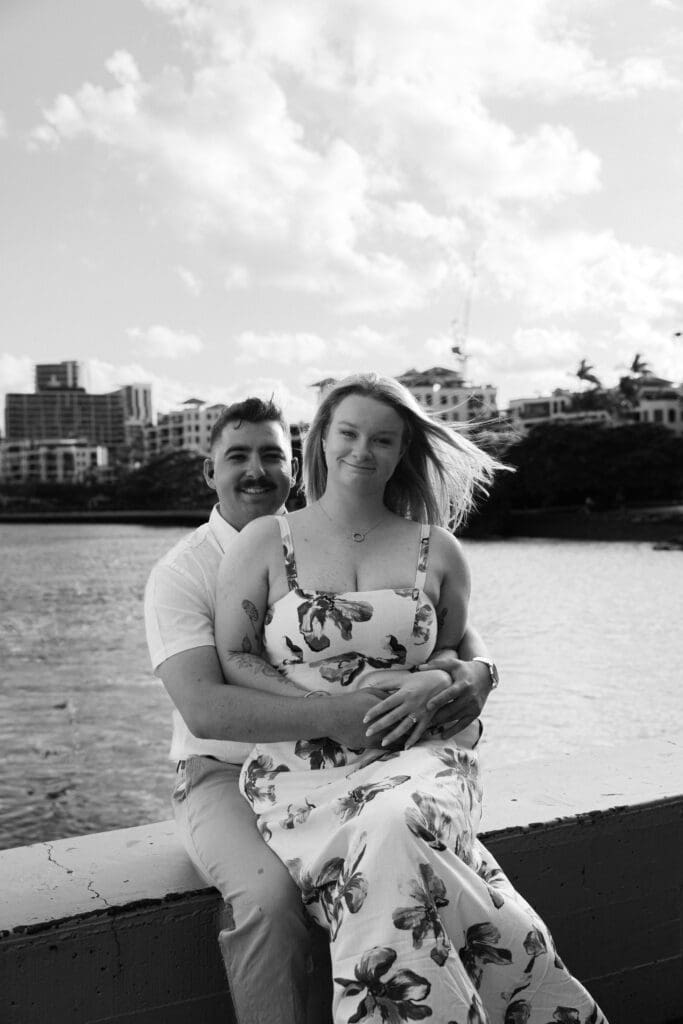 Black and white engagement photo of an engaged couple sitting by the river in Newstead, Brisbane, captured in a relaxed documentary style by a Brisbane wedding photographer.