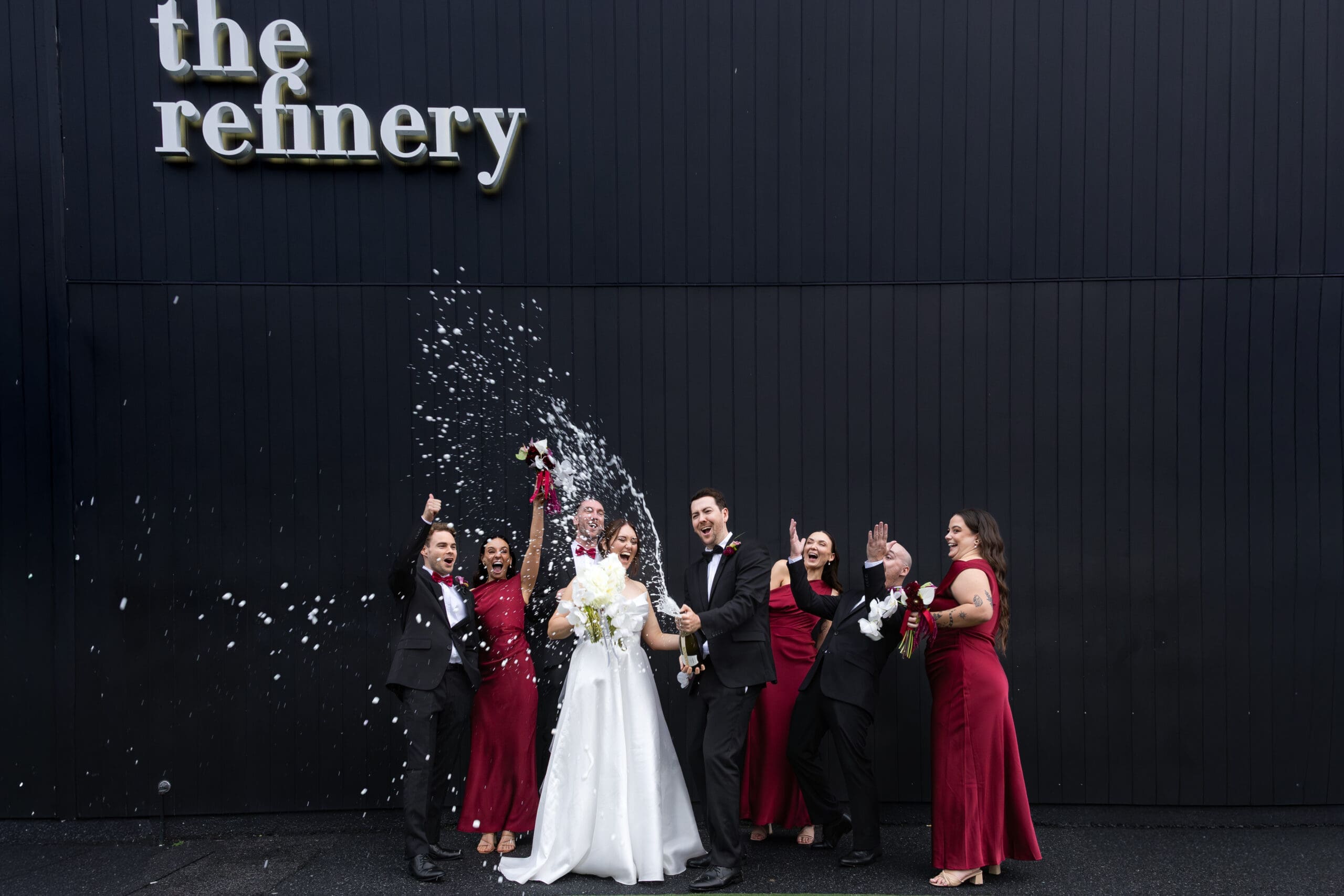 Bride and groom celebrating with their wedding party outside The Refinery wedding venue as champagne sprays into the air during a joyful group photo.